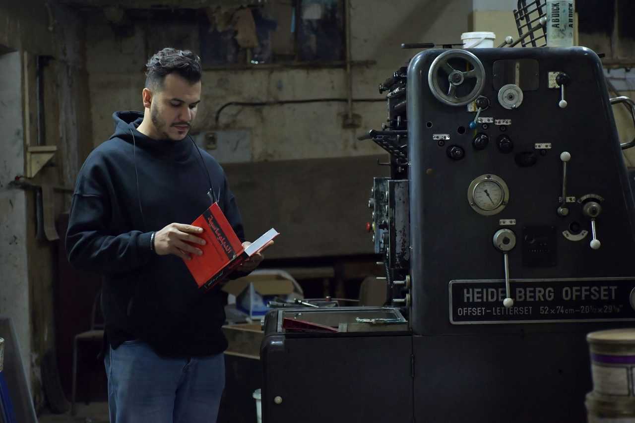 A man in a black hoodie reads by an industrial printing machine in Benghazi, Libya.